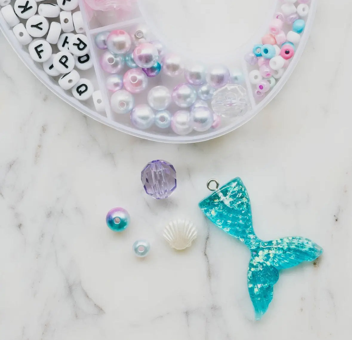 Aquamarine mermaid jewelry kit featuring alphabet beads, shell beads, iridescent star and heart beads, a mermaid tail charm, and pink and clear string. Perfect for creating your own bracelets or necklaces. Made by women-owned small STACKS. Warning: small parts.