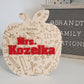 A wooden apple-shaped teacher sign engraved with doodles of school-related items, such as pencils, rulers, and hearts, along with text like 'teaching' and 'ABC.' The center prominently features the name 'Mrs. Kozelka' in bold red letters, perfect as a personalized teacher gift.
