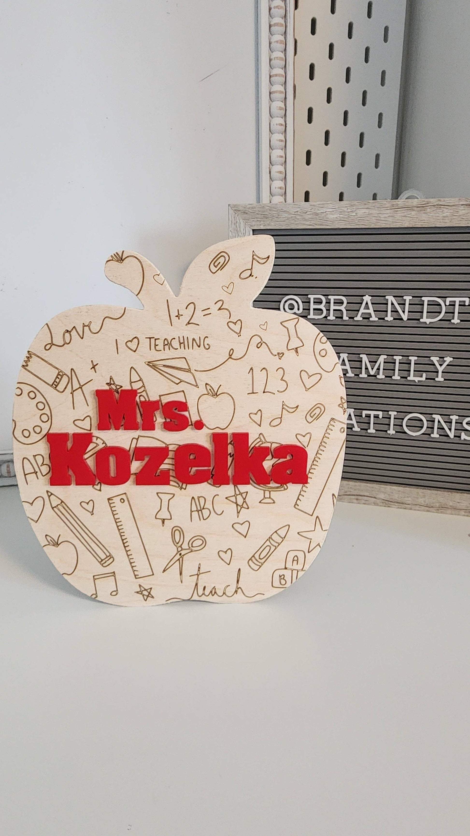 A wooden apple-shaped teacher sign engraved with doodles of school-related items, such as pencils, rulers, and hearts, along with text like 'teaching' and 'ABC.' The center prominently features the name 'Mrs. Kozelka' in bold red letters, perfect as a personalized teacher gift.
