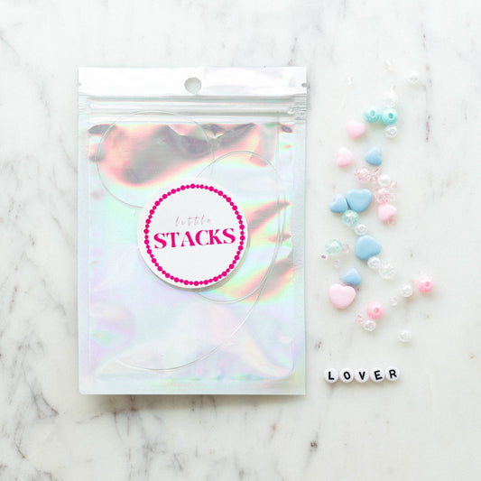 DIY Swiftie friendship bracelet kit featuring light pink and blue heart beads, glitter beads, and clear string. Comes in a silver bag with 'Lover' theme, perfect for creating two custom bracelets. Ideal for Taylor Swift fans and crafting enthusiasts.