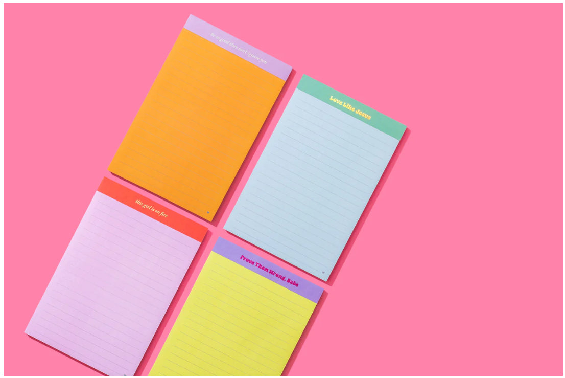 Assortment of colorful motivational notepads on a pink background, featuring phrases like 'Prove Them Wrong, Babe,' 'Love Like Jesus,' 'Believe in Her,' and 'Be Strong, Be Courageous.' Each notepad comes in different colors including yellow, green, pink, and purple, ideal for inspiring notes and personal reflections.