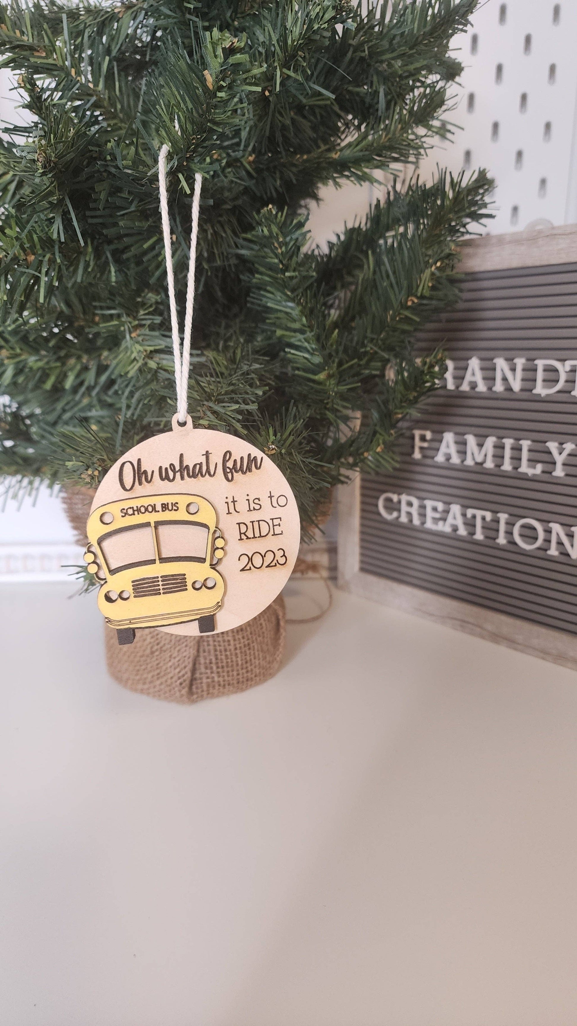 A festive Christmas tree ornament featuring a wooden design with a yellow school bus and the text 'Oh what fun it is to ride 2023.' The ornament hangs on a small Christmas tree with a burlap base, adding a personalized touch to holiday decor.
